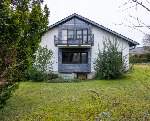 Seitenansicht Haus mit geschiefertem Bereich und Balkon, davon Büsche, Wiese