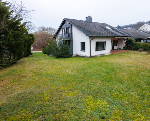 Blick aus dem Garten seitlich auf das Haus, man sieht viel Rasen im Vordergrund, links mehrere Nadelbäume und dann das Haus seitlich mit Balkon und Loggia.