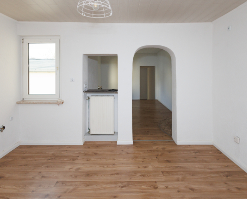 Zimmer mit weißen Wänden und Torbogen rechts, daneben Durchreiche und Fenster, Boden helles Holz