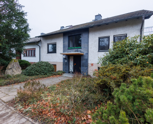 Immobilie mit leicht seitlicher Ansicht von rechts, rechts sind einige Sträucher mit Herbstzustand, dann Weg zum Haus daneben wieder Sträucher und ein hoher Nadelbaum. Immobilie leicht nach hinten gesetzt