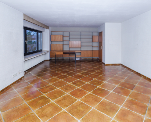 großes Wohnzimmer mit großen braunen Bodenfliesen, alles andere hell, an der Rückwand zeitlose Regalwand mit Schreibtisch davor, zwei Zimmertüren rechts, links ein großes Fenster